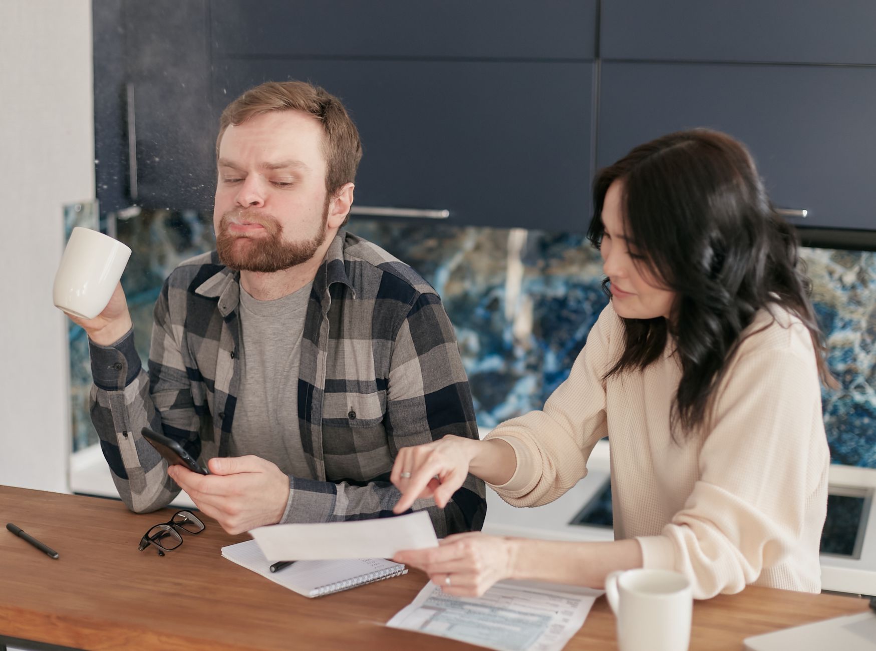 A couple at a kitchen counter with printed paperwork, a notebook and a phone, going through household bills together.