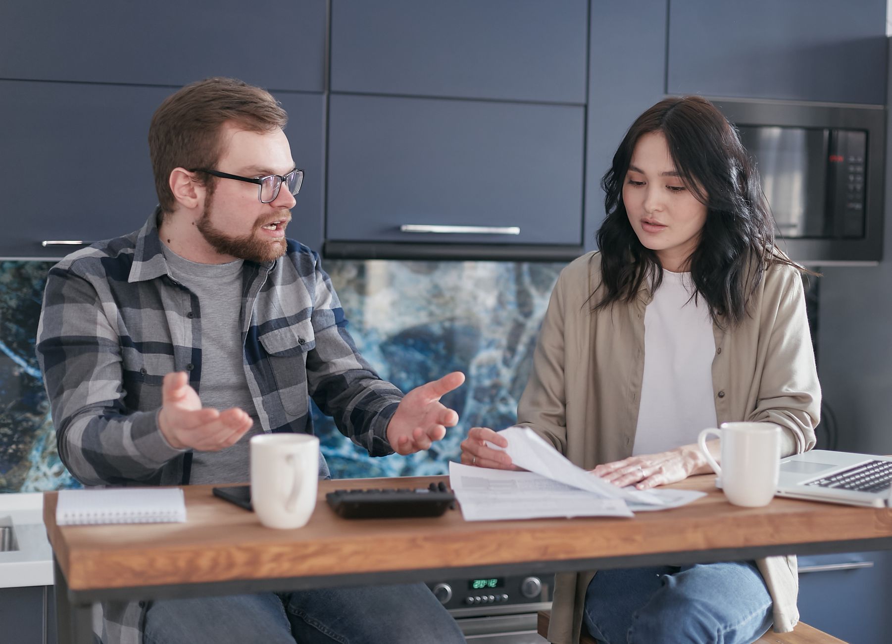 A man and a woman at a kitchen counter go through printed paperwork together, with mugs, a notebook and a laptop beside them.